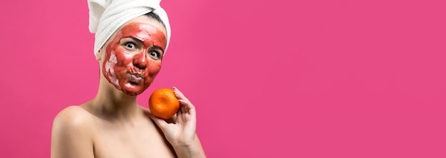 Piękno portret kobiety w białym ręczniku na głowie z czerwoną odżywczą maską na twarzy Pielęgnacja skóry oczyszczająca eko organiczny kosmetyczny spa relaks koncepcja Dziewczyna stoi z plecami trzymając pomarańczową mandarynkę