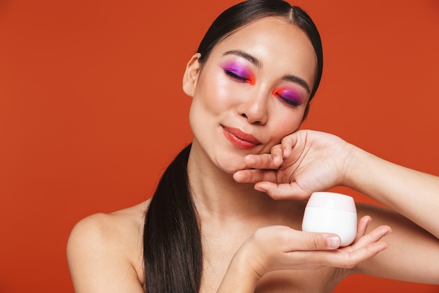 Piękno portret szczęśliwej młodej topless azjatyckiej kobiety z brunetką w jasnym makijażu, stojącej odizolowanej na czerwono, pokazującej butelkę z kremem do twarzy