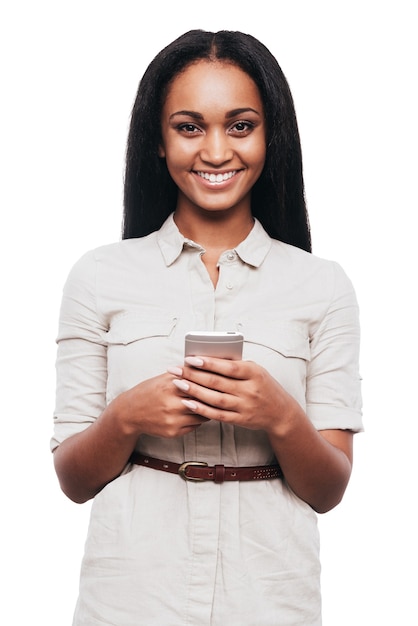 Piękno z inteligentnym telefonem. Piękna młoda afrykańska kobieta trzyma telefon komórkowy i uśmiecha się stojąc na białym tle
