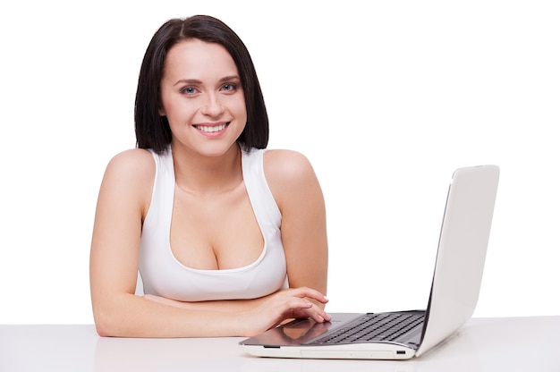Piękno z laptopem. Atrakcyjna młoda kobieta z pięknym dekoltem pracuje na laptopie i uśmiecha się siedząc przy stole i na białym tle