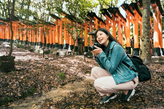 piękny azjatycki backpacker klęcząc w lesie przyrody w japońskiej świątyni trzymając profesjonalny aparat robienia zdjęć. młoda dziewczyna fotograf w inari sanktuarium kioto japonia radośnie się uśmiechając.