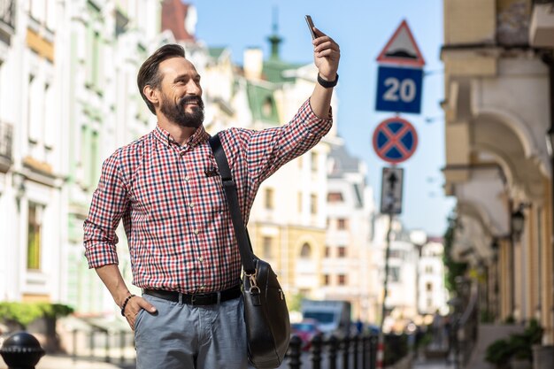 Piękny budynek. Radosny przystojny mężczyzna spaceruje ulicą podczas robienia zdjęć