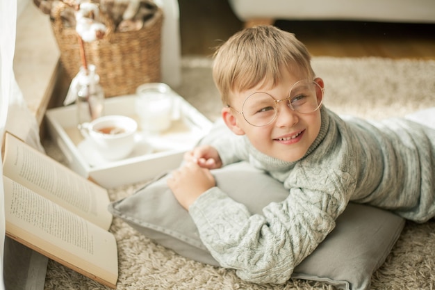 Piękny chłopczyk w swetrze z dzianiny czyta przy oknie z kubkiem gorącej herbaty. Przytulny. Jesień. Jesienny nastrój.