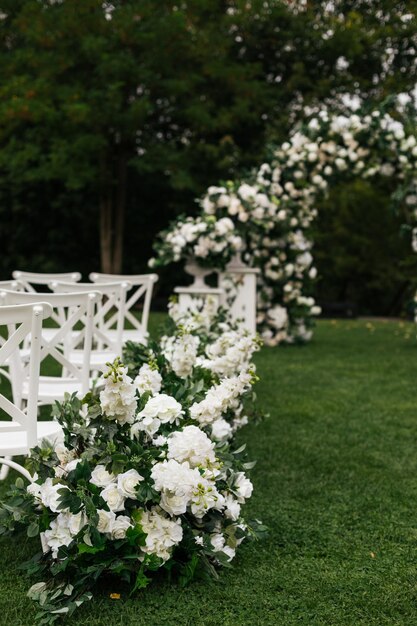 Piękny i stylowy łuk ślubny ozdobiony różnymi świeżymi białymi kwiatami z białymi krzesłami