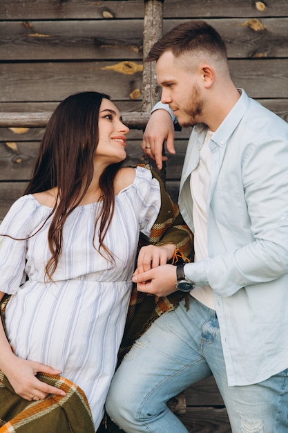 Piękny kobieta w ciąży i jej mąż pozuje blisko drewnianego budynku. Stylowa szczęśliwa para kocha się. Moda i rustykalny styl.