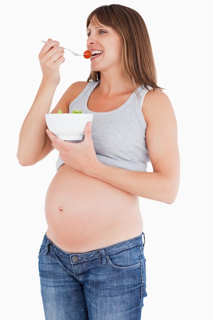 Piękny kobieta w ciąży je czereśniowego pomidoru podczas gdy trzymający puchar sałatka
