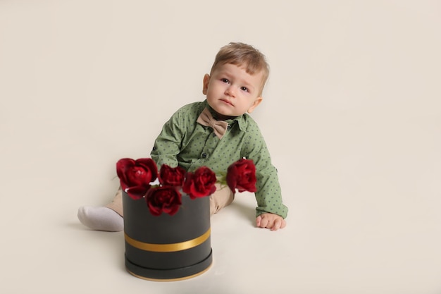 piękny mały chłopiec w zielonej koszuli siedzi na białym tle obok pudełka z czerwonymi różami
