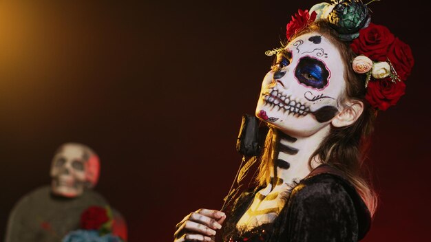 Piękny model ubrany w kostium śmierci i wieniec kwiatów na tle studia, działając jak święty santa muerte. La cavalera catrina dama zmarłych ze sztuką ciała czaszki, kultura meksykańska. Strzał z ręki.