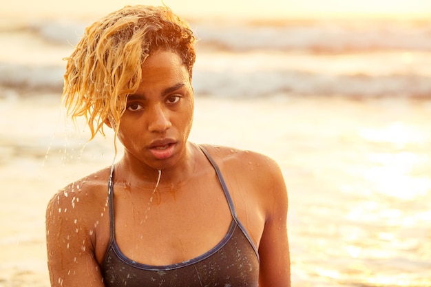 Piękny portret afroamerykańskiej surferki z mokrymi afro blond kręconymi włosami w bikini o zachodzie słońca na plaży