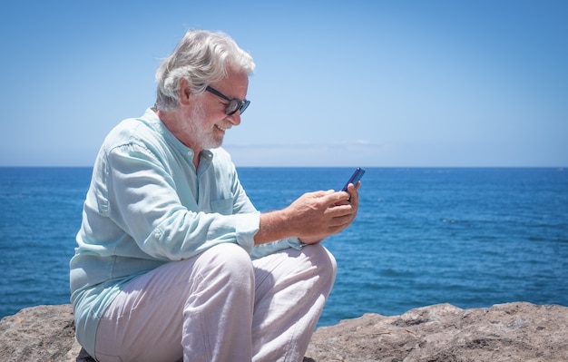 Piękny starszy mężczyzna siedzący na świeżym powietrzu nad morzem podczas korzystania z telefonu komórkowego, ciesząc się światłem słonecznym