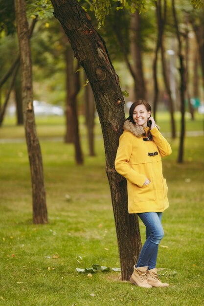 Piękny szczęśliwy kaukaski młody uśmiechający się brązowe włosy kobieta w żółtym płaszczu, dżinsy, buty w zielonym lesie. Moda modelki ze złotymi liśćmi jesienią stojąc i spacerując w parku wczesną jesienią na zewnątrz.