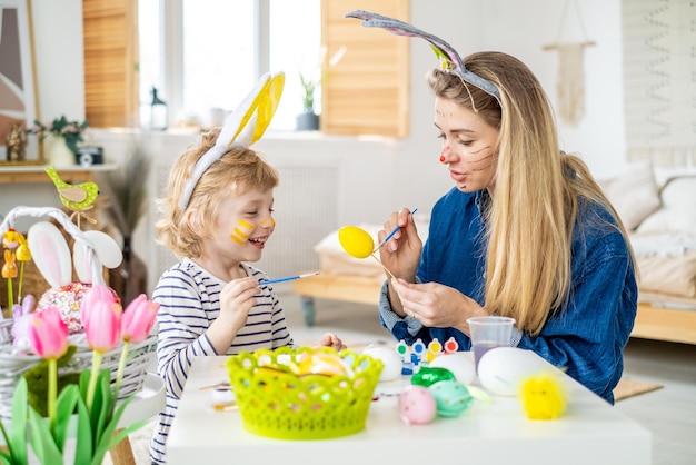 Piękny szczęśliwy syn i matka w opaskach na głowę z króliczymi uszami dekorują jajka pędzlem i jasnymi farbami przygotowują się do świętowania Wielkanocy baw się dobrze w domu