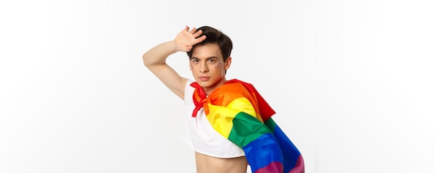 Piękny wesoły z brokatem na twarzy noszący crop top i tęczową flagę lgbt pozuje na białym b