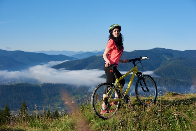 Piękny żeński rowerzysta z żółtym bicyklem w górach w ranku. Mgliste góry, lasy. Aktywność na świeżym powietrzu