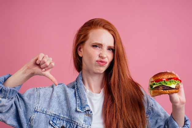 Piękny źle redhaired rudy imbir wegetariańska kobieta trzyma duży cheeseburger z serem, kotletem wołowym i sałatą z pomidorów w studio różowym tle.