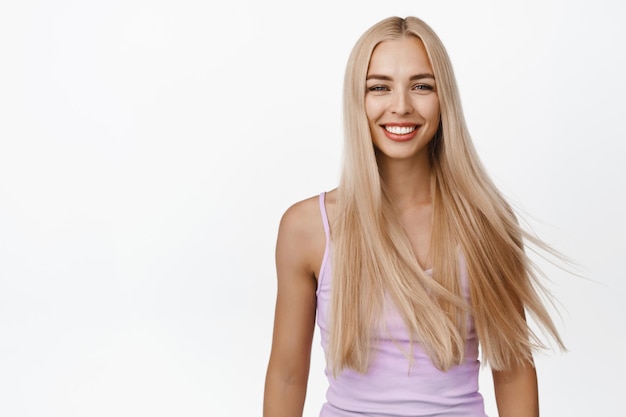 Zdjęcie pielęgnacja włosów i dobre samopoczucie piękna blond dziewczyna ze świecącymi zdrowymi długimi włosami uśmiecha się szczęśliwa do kamery stojącej w podkoszulku na białym tle