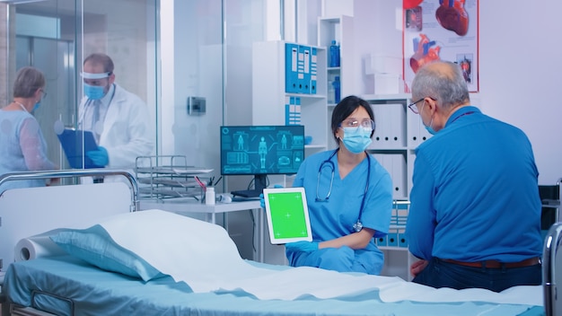 Pielęgniarka prezentuje tablet z zielonym ekranem pacjentowi w nowoczesnym prywatnym szpitalu. Izolowany ekran zastępczy chromatycznej makiety na gadżecie dla Twojej aplikacji, tekstu, wideo lub zasobów cyfrowych. Łatwe kluczowanie medycyna