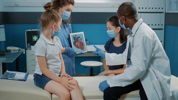 Pielęgniarka trzymająca obraz kardiologiczny i medyk wyjaśniający stan serca podczas badania. Asystent i lekarz analizujący diagnozę sercowo-naczyniową z dzieckiem i matką podczas pandemii Covid 19.