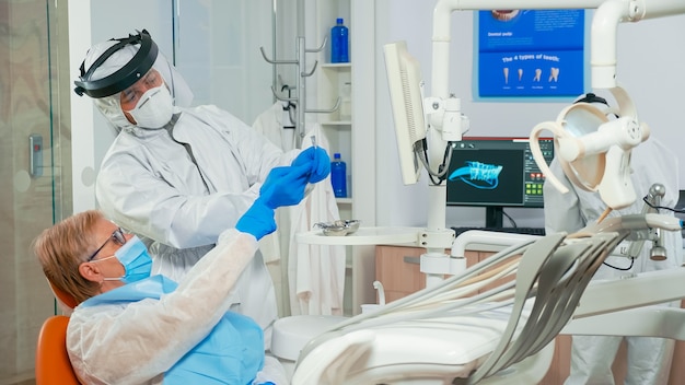 Pielęgniarka w kombinezonie pomaga lekarzowi z RTG jamy ustnej pacjenta podczas badania w pandemii koronawirusa. Zespół medyczny noszący maskę na twarz, rękawiczki, wyjaśniający radiografię za pomocą wyświetlacza monitora;