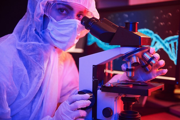 Pielęgniarka w masce i białym mundurze siedzi w oświetlonym neonami laboratorium z komputerem, mikroskopem i sprzętem medycznym poszukuje szczepionki przeciwko koronawirusowi