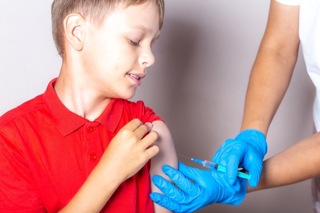 Pielęgniarka w niebieskich rękawiczkach wstrzykuje szczepionkę przez strzykawkę w ramię chłopca