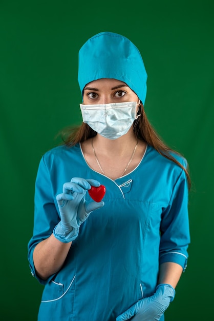 Pielęgniarka w niebieskim mundurze z rękawiczkami z maską trzymająca małe czerwone serce w szpitalu na sali operacyjnej