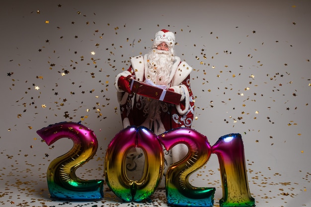 Pień fotografia wesołego ojca mrozu w świątecznym ubraniu z długą białą brodą wyciągając ramiona i uśmiechając się do kamery pod latającym złotym konfetti. Na białym tle na szarym tle.