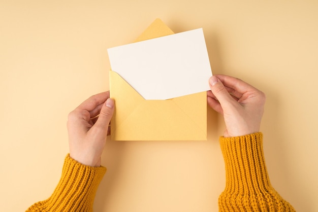 Pierwsza osoba zdjęcie widoku z góry kobiecych rąk w żółtym swetrze trzymającym otwartą pastelową żółtą kopertę z białą kartką na na białym tle jasnopomarańczowym z copyspace