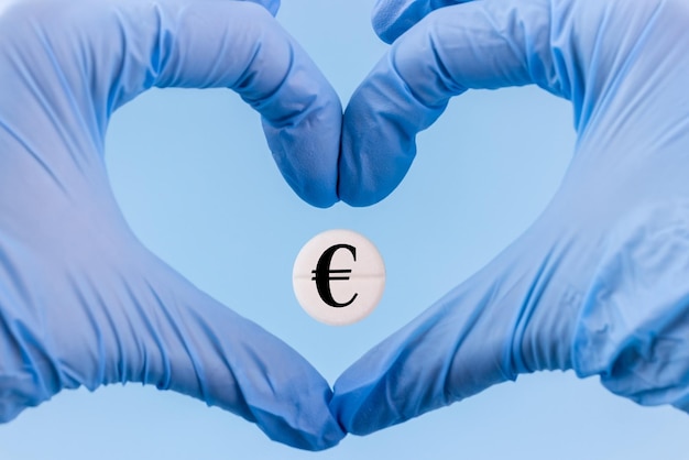 Pigułka ze znakiem waluty Euro w sercu wykonanym z rąk w rękawiczkach medycznych na niebieskim tle Wzrost cen leków w Europie Drogie leczenie