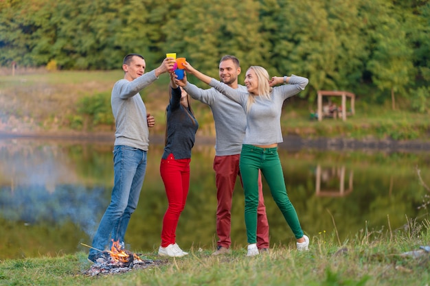 Piknik z przyjaciółmi przy jeziorze w pobliżu ogniska. Firma przyjaciele ma podwyżki natury pyknicznego tło. Turystów relaks w czasie picia. Letni piknik. Zabawa z przyjaciółmi