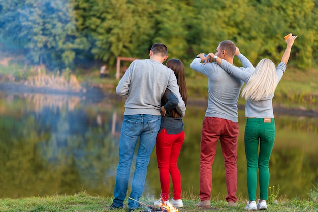 Piknik z przyjaciółmi przy jeziorze w pobliżu ogniska. Firma przyjaciele ma podwyżki natury pyknicznego tło. Turystów relaks w czasie picia. Letni piknik. Zabawa z przyjaciółmi