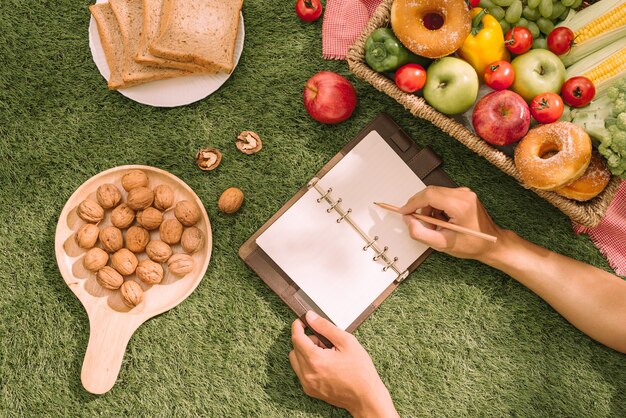 Piknikowy wiklinowy kosz z jedzeniem, chlebem, owocami i sokiem pomarańczowym na czerwono-białej szachownicy w polu z zielonym tłem przyrody. Koncepcja pikniku.