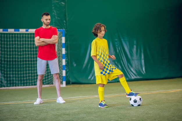 Piłka nożna. Słodki chłopak w żółtym mundurze gra w piłkę nożną, jego trener obserwuje go