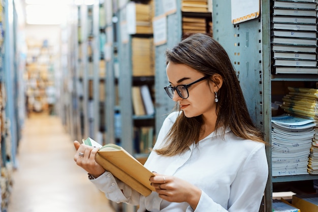Zdjęcie pilna studentka w białej bluzce i okularach czytająca książkę w bibliotece