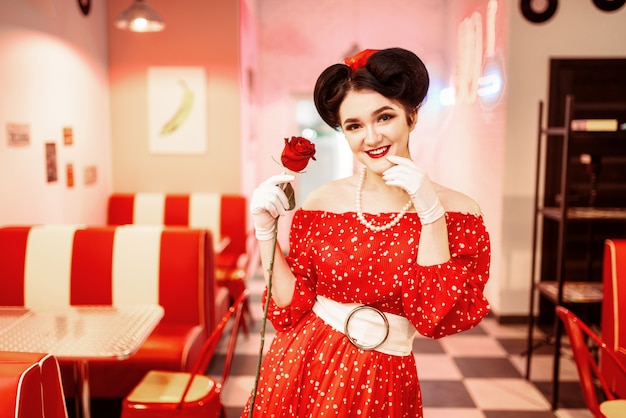 pin up kobieta z makijażem trzyma czerwoną różę, sukienka w kropki, styl vintage. Wnętrze kawiarni retro z podłogą szachownicy