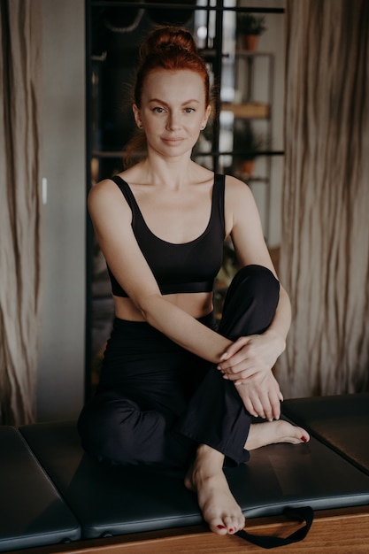 Pionowe ujęcie fit młoda wysportowana kobieta siedzi z rękami wokół nóg w pozycji relaksacyjnej po pilates lub treningu fitness na trapezie w siłowni. Pojęcie zdrowego stylu życia i sportu