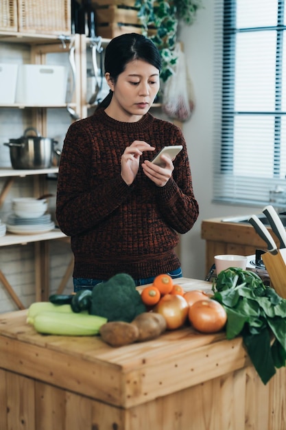 pionowe ujęcie gospodyni azjatycka zamawia świeże warzywa na swoim smartfonie podczas zakupów spożywczych online przy stole kuchennym w jasnym wnętrzu domu.