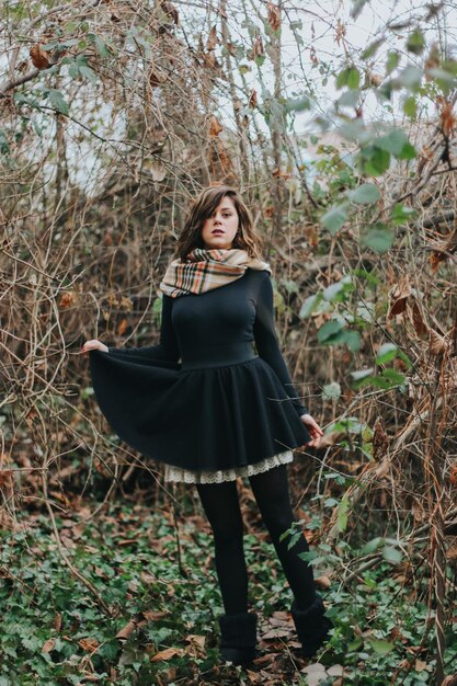 Pionowe ujęcie kaukaskiej kobiety ubranej w czarną sukienkę i szalik w lesie jesienią