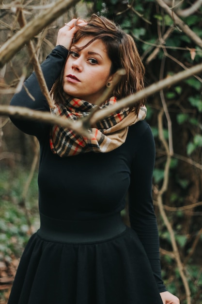 Pionowe ujęcie kaukaskiej kobiety ubranej w czarną sukienkę i szalik w lesie jesienią