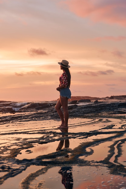 Pionowe ujęcie kobiety w szortach i kapeluszu stojącej na plaży podczas pięknego zachodu słońca