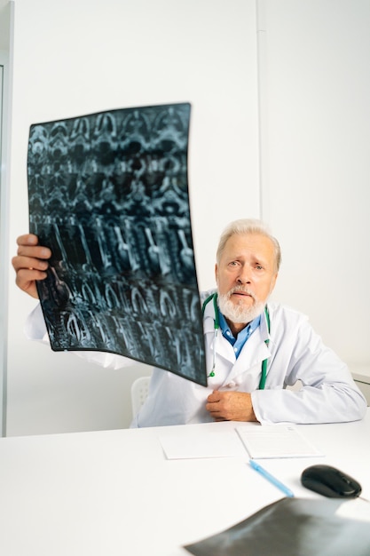 Zdjęcie pionowe ujęcie poważnego dojrzałego dorosłego mężczyzny lekarza trzymającego obraz kręgosłupa mri pacjenta siedzącego przy biurku w pokoju gabinetu lekarskiego, patrzącego na kamerę, myślącego o diagnozie, wyrażającej zaniepokojenie chorobą