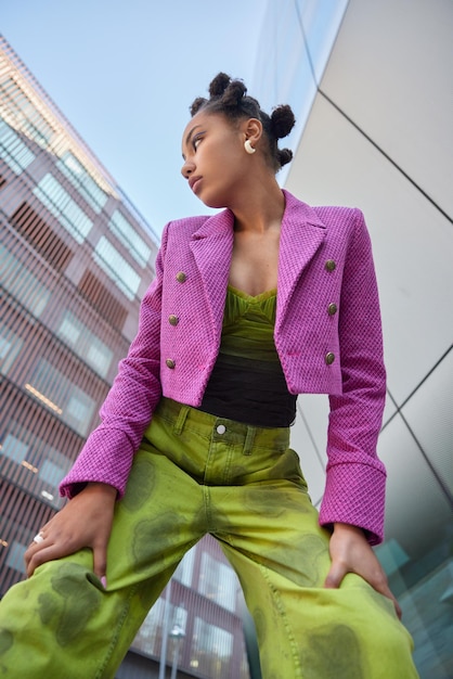 Pionowe ujęcie stylowej kobiety ubranej w różową kurtkę i zielone spodnie, pochylającej się na kolanach, skupionych w pozach na tle miejskich budynków w mieście coś zauważa. Koncepcja mody i stylu życia ludzi