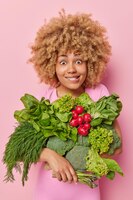 Zdjęcie pionowe ujęcie zaskoczonej kobiety z ugryzionymi kręconymi włosami. wargi niosące świeże zielone surowe warzywa z ugryzień szklarniowych pozują na różowym tle pochodzą z rynku wegańskie składniki żywności