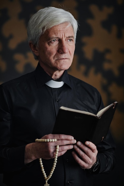 Pionowo spokojny starszy ksiądz trzymający Biblię w dramatycznym oświetleniu