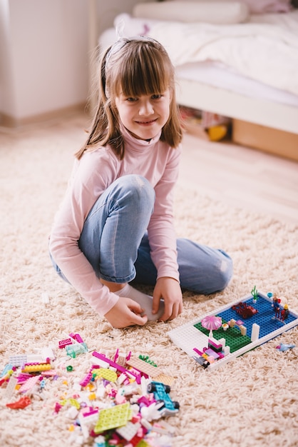 Pionowo widok dosyć uśmiechnięta mała berbeć dziewczyny obsiadanie na dywanie jej pokój i patrzeć kamerę podczas gdy bawić się z plastikowymi zabawkami.