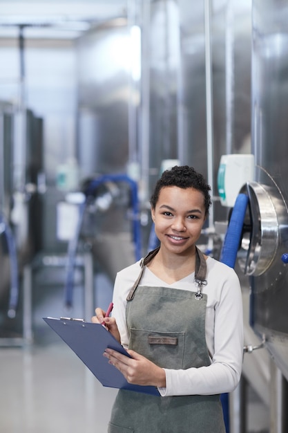 Pionowy pas w górę portret młodej afroamerykańskiej kobiety cieszącej się pracą w fabryce przemysłowej i uśmiechającej się do aparatu trzymającego schowek, kopia przestrzeń