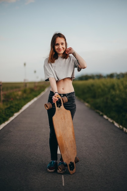 Pionowy plakat portret młodej kobiety z tatuażem opierając się na longboardzie patrzy w kamerę kobieta stojąca na środku drogi