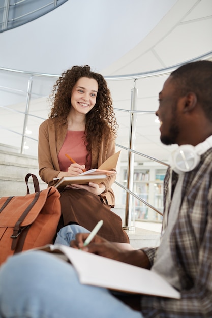 Pionowy portret dwóch studentów rozmawiających na schodach w college'u i pracujących nad pracą domową, skup się na młodej, kręconej, uśmiechniętej wesoło młodej kobiecie