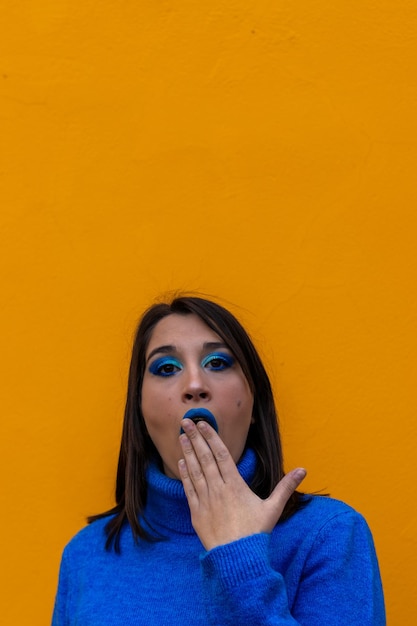 Pionowy portret kaukaskiej kobiety ubranej i umalowanej na niebiesko ze zdziwionym gestem z ręką w ustach na żółtym tle