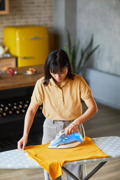 Pionowy portret młodej Azjatki prasującej ubrania w domu i wykonującej prace domowe
